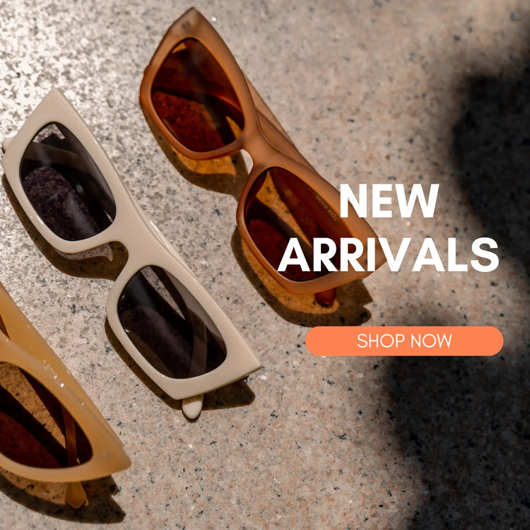 Three stylish brown and beige sunglasses placed on a sunlit concrete floor. The text 'NEW ARRIVALS' is prominently displayed in white, with a call-to-action button labeled 'SHOP NOW' in orange.